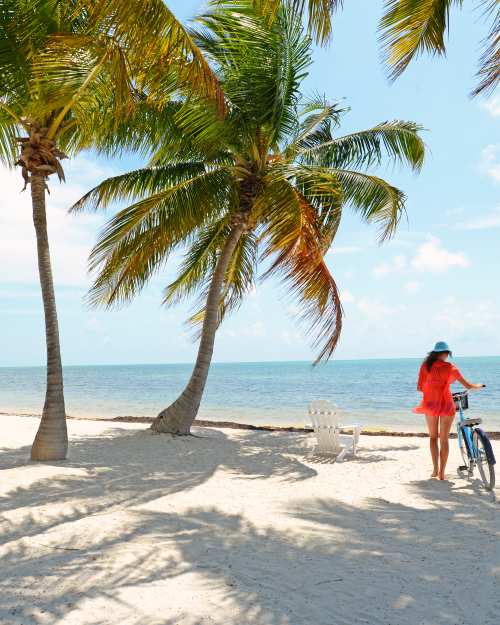 Girl riding blue bike on Florida Keys beach with palm trees and white adirondack chair